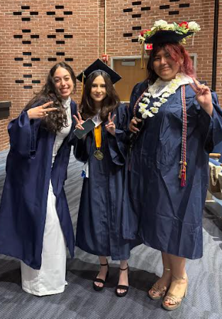 Three women in blue gowns and caps pose for a photo in a room with a brick wall.