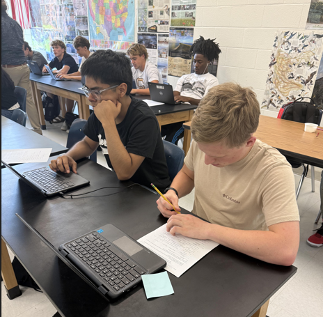 Two students are working at a table in a classroom. One is using a laptop, and the other is writing.
