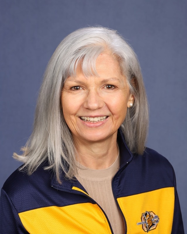 An older woman with white hair smiles. She wears a blue and yellow jacket with a logo on the left chest area.