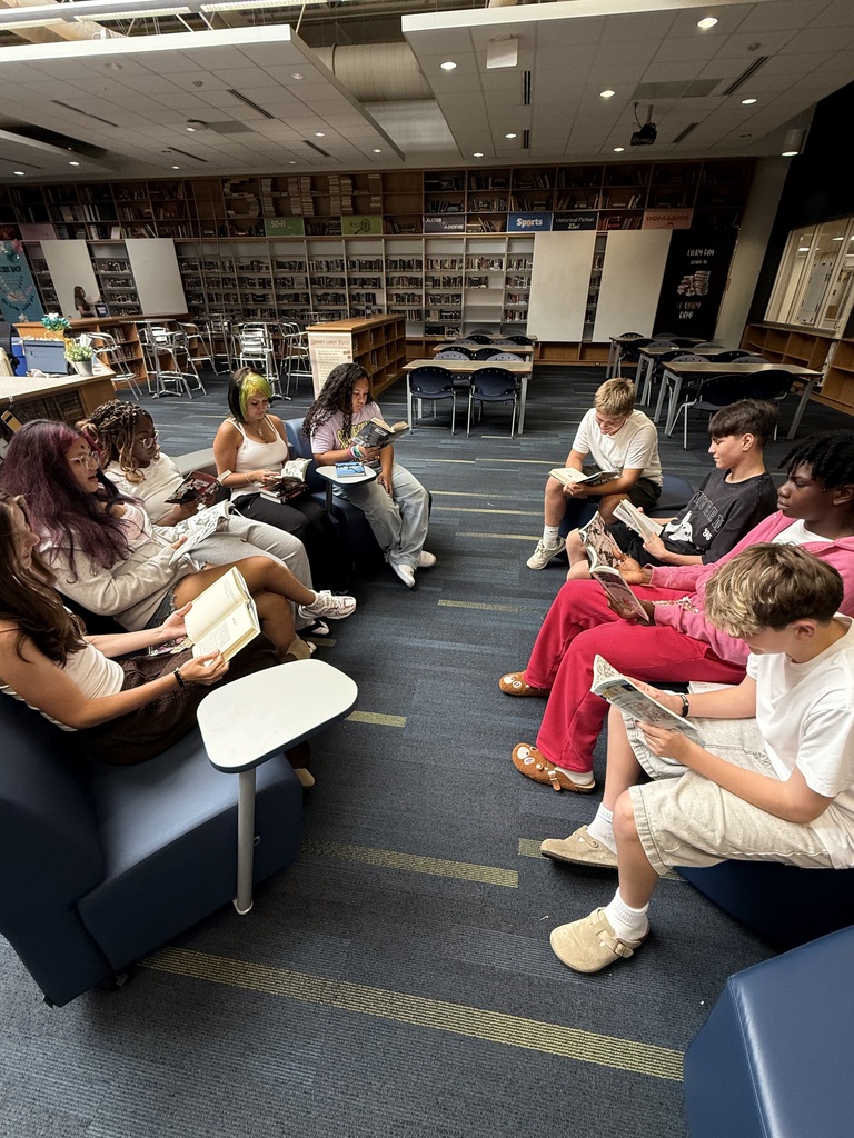 A group of students sit around reading books in a library, with bookshelves and tables in the background.