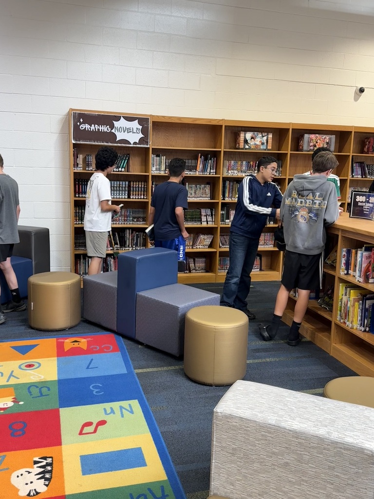 People in a library with bookshelves. One person is standing next to a colorful rug.