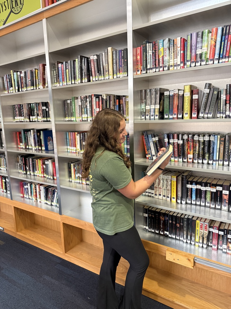 Woman standing in library with books in hand, shelves filled with books behind her.