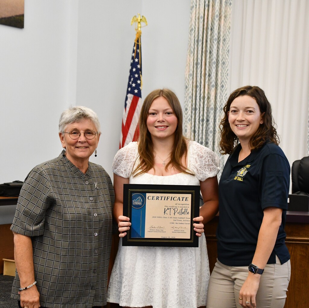 Three women, one with a certificate, stand in a room. Two wear polo shirts; one wears a dress.