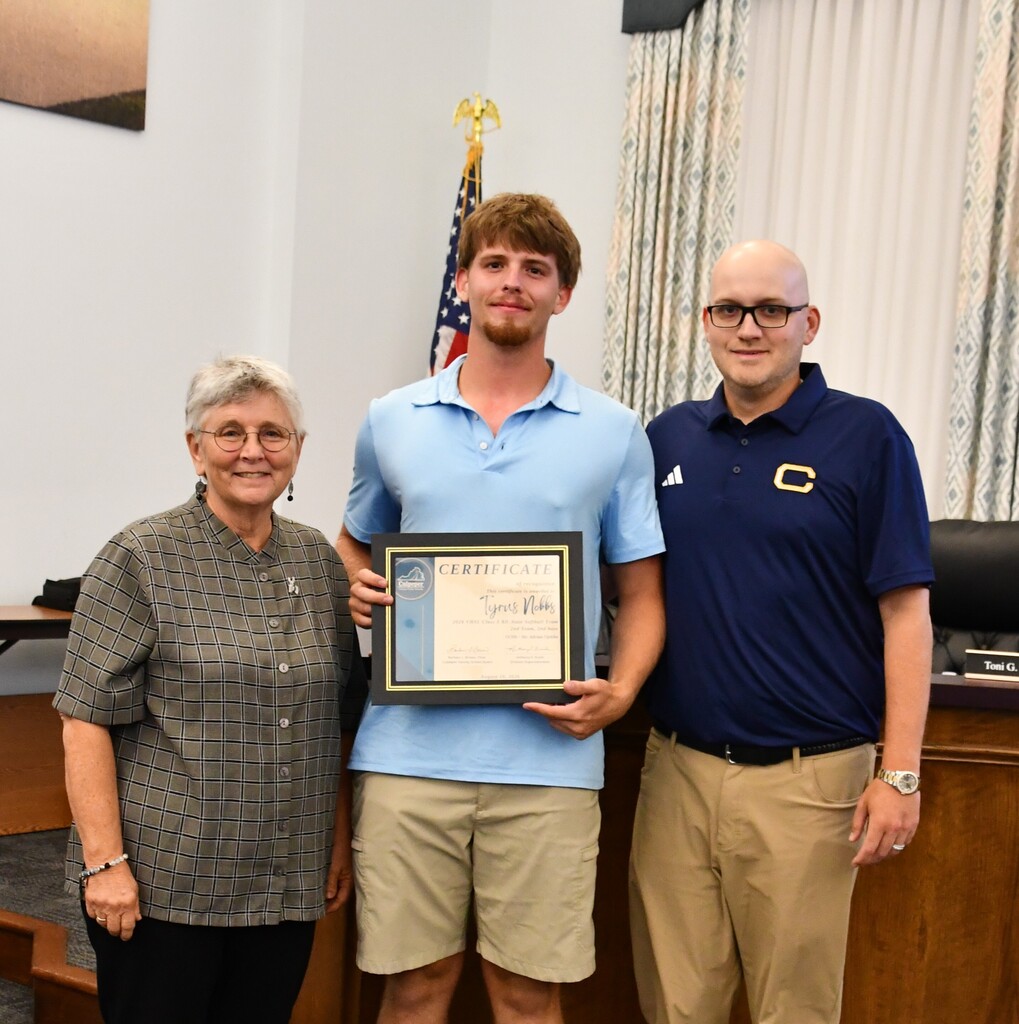 Three individuals, one holding an award, stand together in a board room. The young man in the middle wears a light blue polo. and stands in between his coach and a School Board memvber