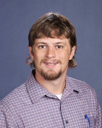 A man with a beard and brown hair wearing a checkered shirt, smiling and looking at the camera.