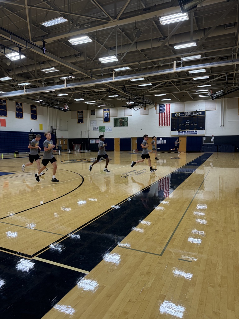 Five people run on a basketball court. The floor has black and wooden markings. The ceiling has several fluorescent lights.