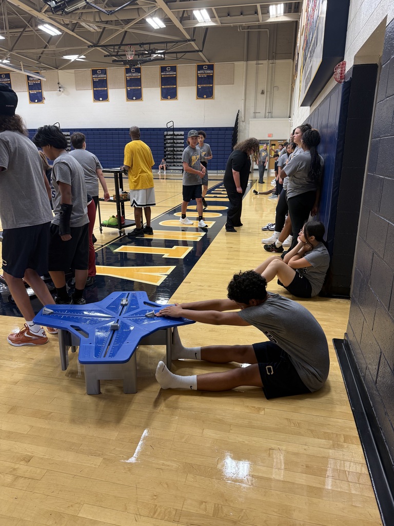 Indoor basketball court with people. Some sit and others stand. A boy kneels to examine a blue object.