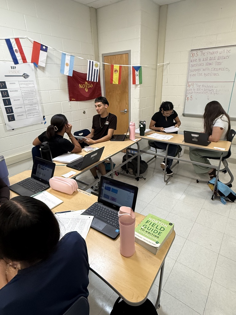 Five students are seated at a desk, using laptops. There are flags hanging on the wall.
