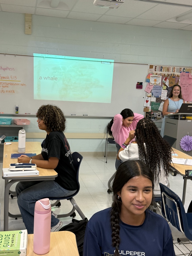 A classroom with students seated at desks. One student sits in front, facing the camera, while others sit behind. A teacher stands near a podium.