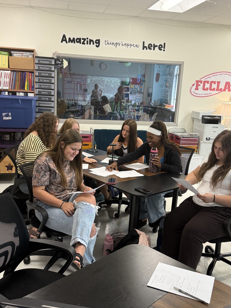 A group of people sits around a table in a classroom, with papers and pens. A banner reads "Amazing things happen here!"