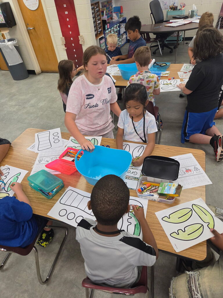 Older students help younger students cut, color, and assemble paper craft pieces at a classroom table.