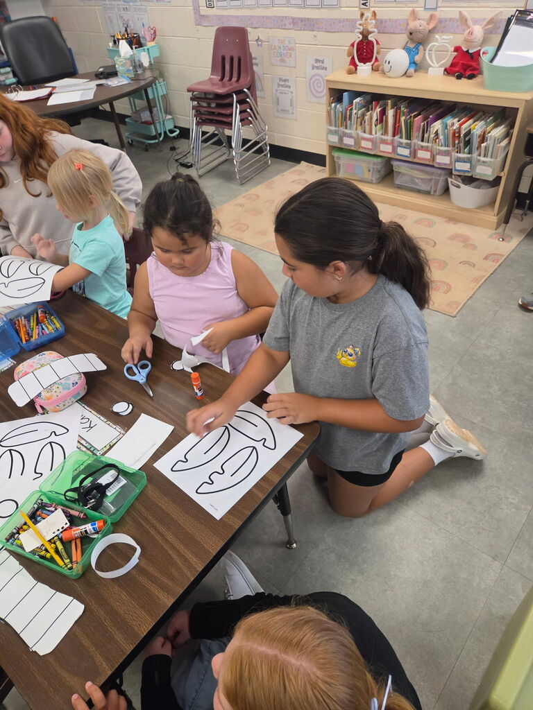 Older students help younger students cut, color, and assemble paper craft pieces at a classroom table.