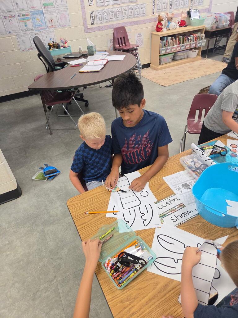 Older students help younger students cut, color, and assemble paper craft pieces at a classroom table.