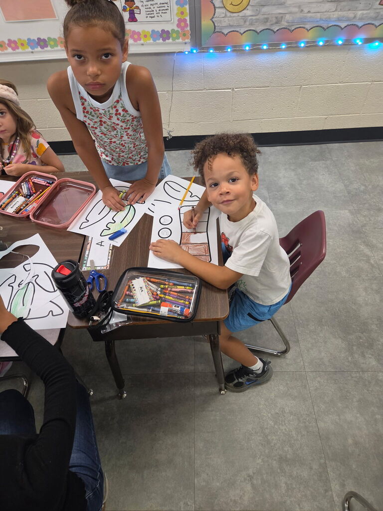 Older students help younger students cut, color, and assemble paper craft pieces at a classroom table.