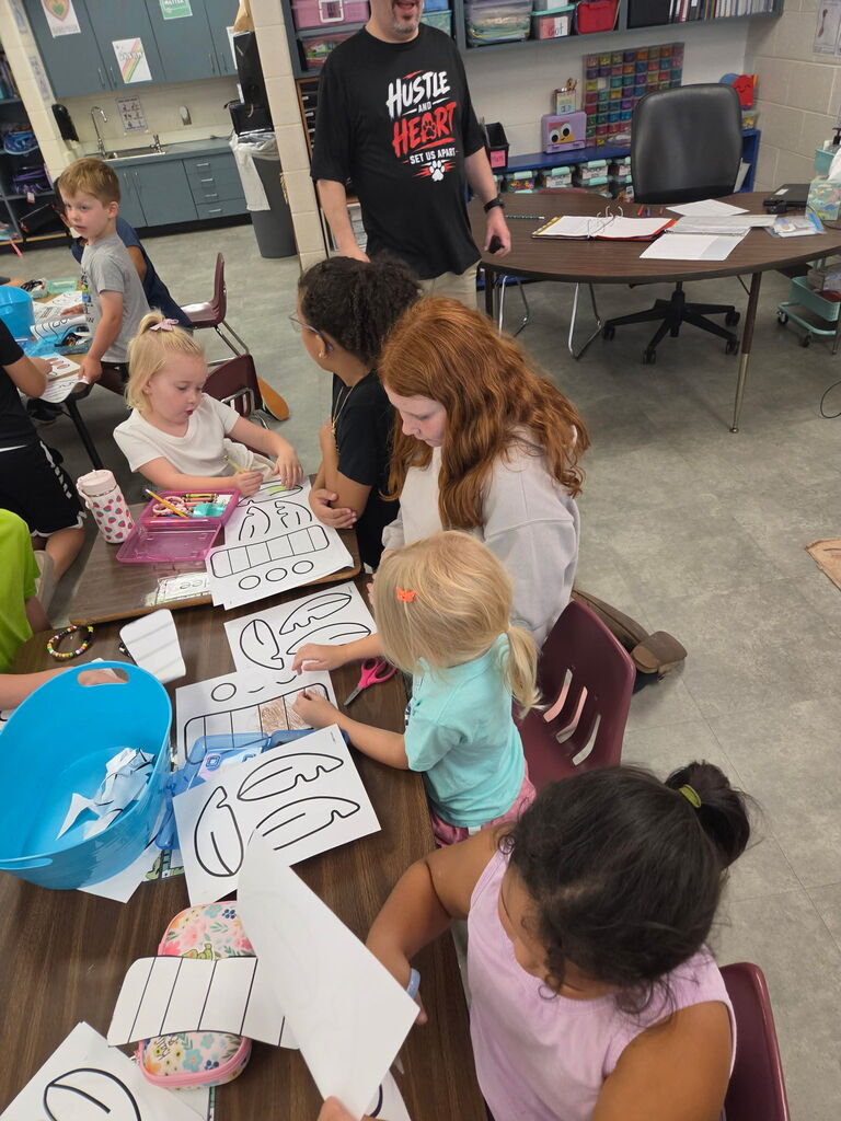 Older students help younger students cut, color, and assemble paper craft pieces at a classroom table.