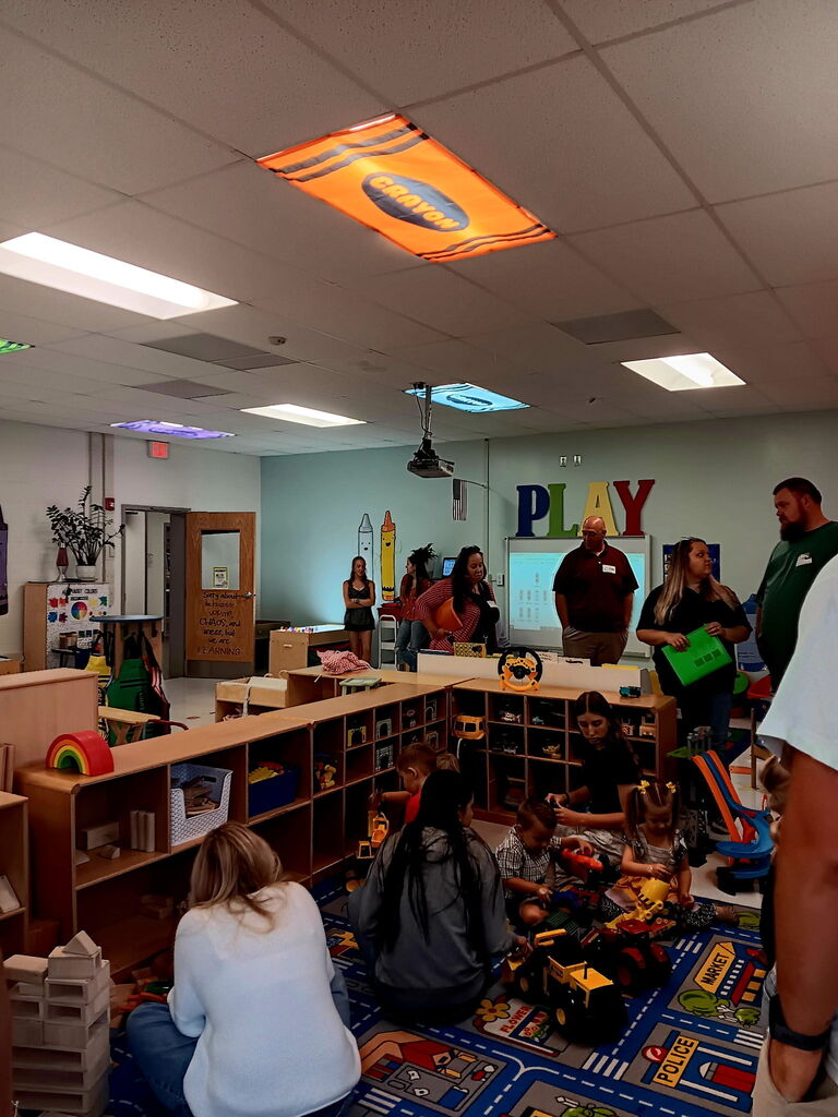 A group of adults and children are gathered in a room with shelves, a carpeted floor, and toys.