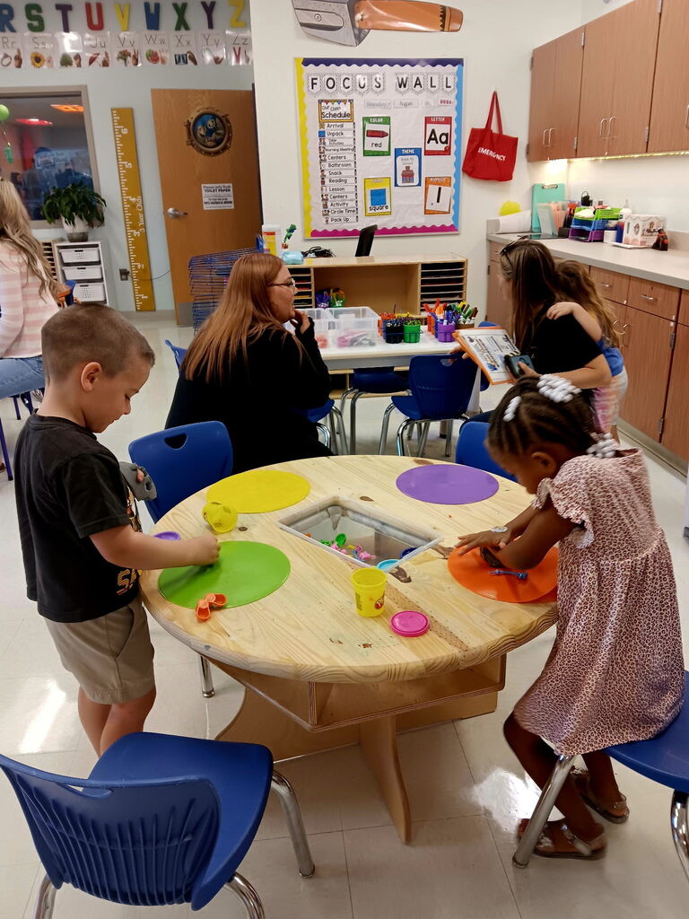 Kids seated around a round table with mats and cups. One child is seated while another stands.