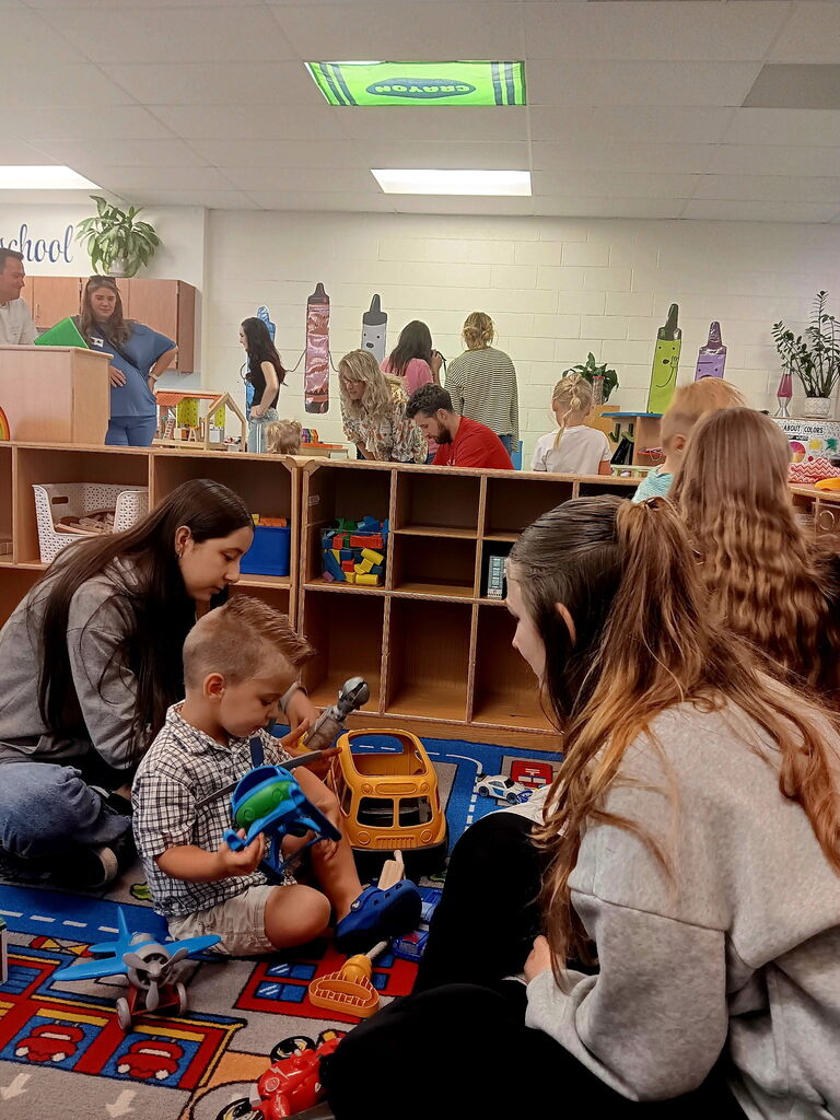 Group of women and children in a play area. A boy plays with a toy truck on the floor.