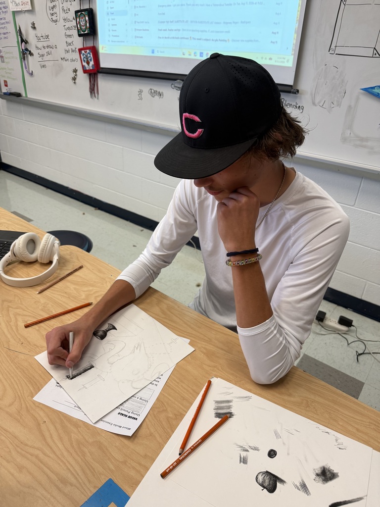 Person seated at a desk, working on drawings with pencils. Background shows a classroom with a whiteboard.