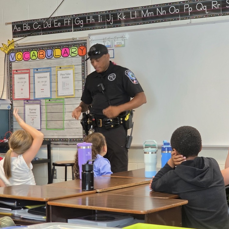Police officer speaks to elementary students seated at desks in a classroom, with one student raising her hand.