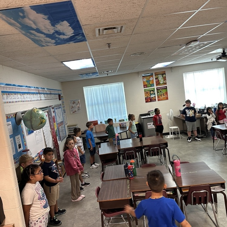 Elementary students stand around the classroom participating in a teacher-led activity.