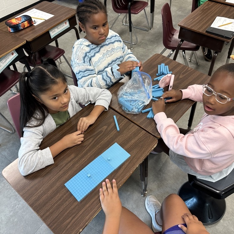 Four elementary students gather around desks and use blue base-ten blocks during a hands-on math activity.