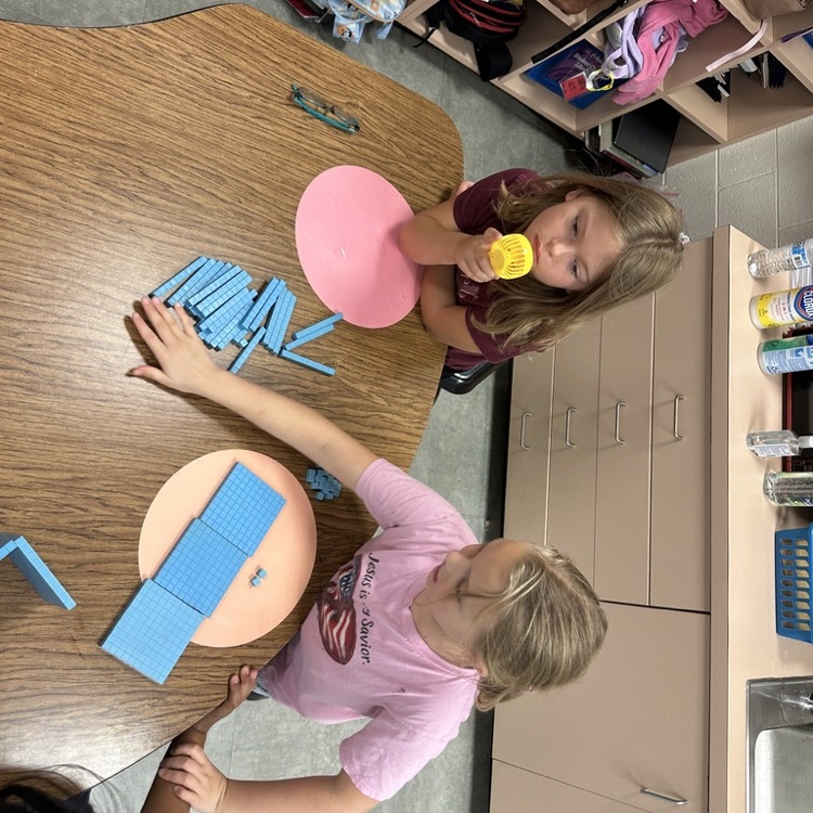 Students work together at a classroom table using blue base-ten blocks and other hands-on math materials.