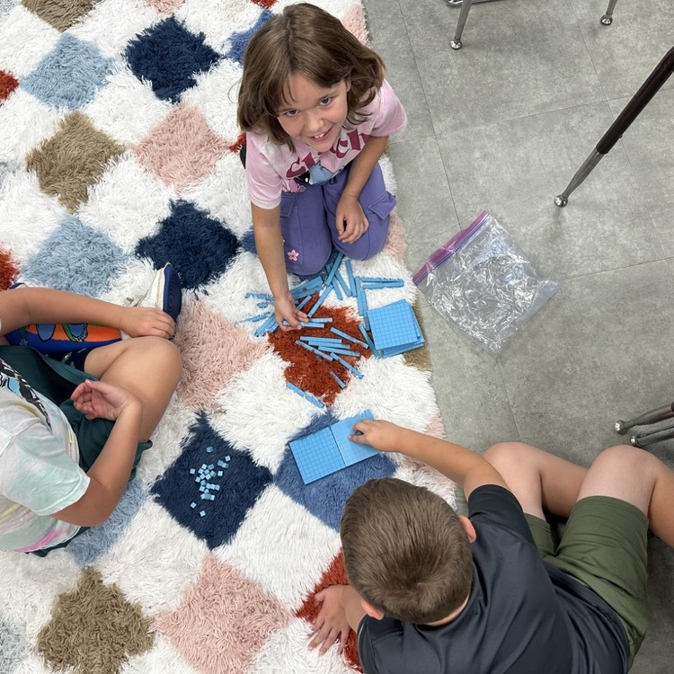 Three elementary students sit on a classroom rug working together with blue base-ten blocks.