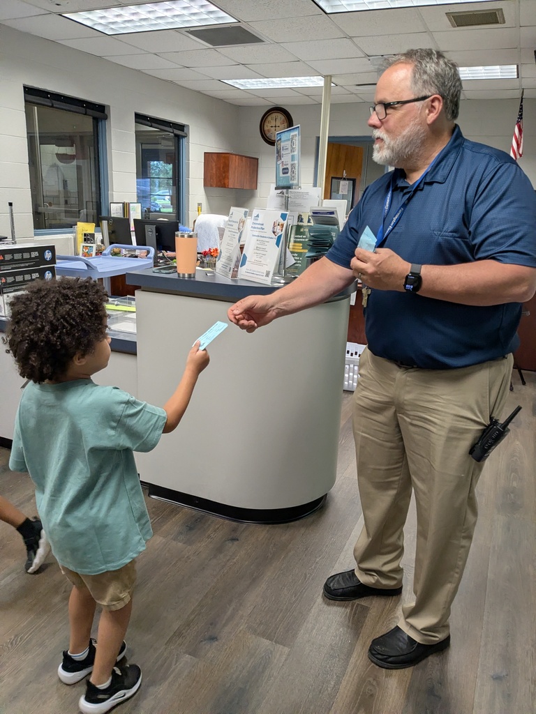 A man in blue and khakis hands a paper to a child in a green shirt. Behind them, a counter holds items and a flag.