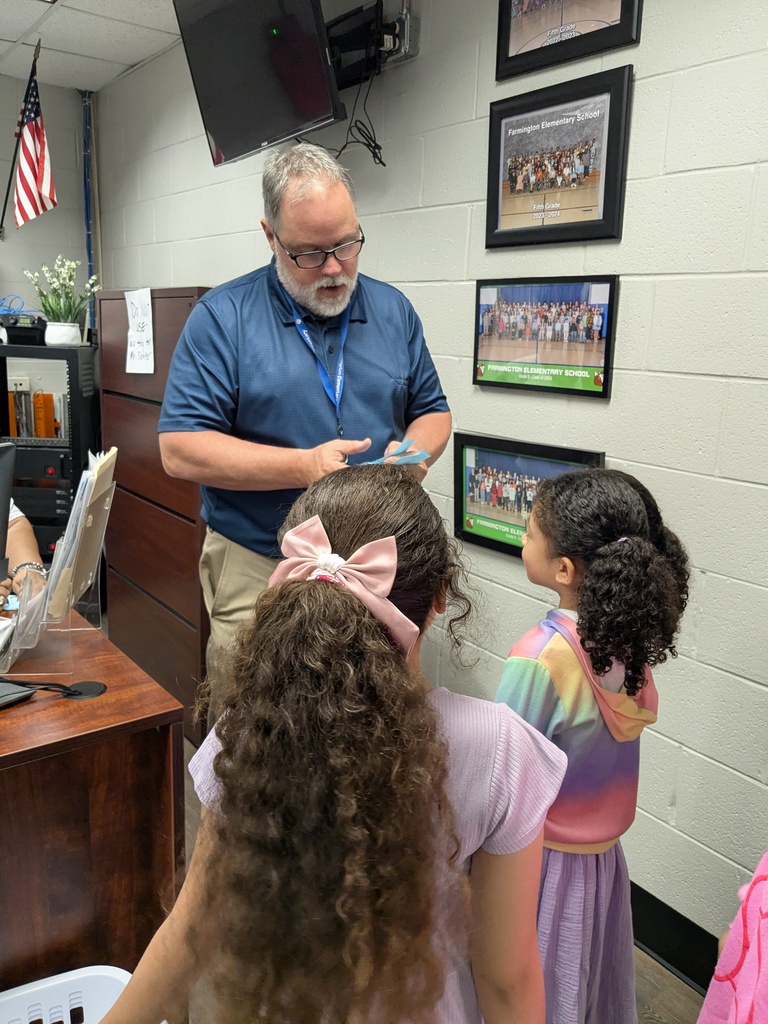 A man in a blue shirt shows something to two young girls with curly hair. A desk, flag, and framed photos are in the background.