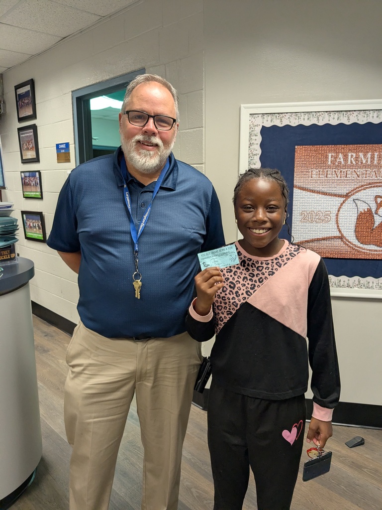 A man with a beard stands next to a girl with a backpack, holding a card. They are in a school hallway.