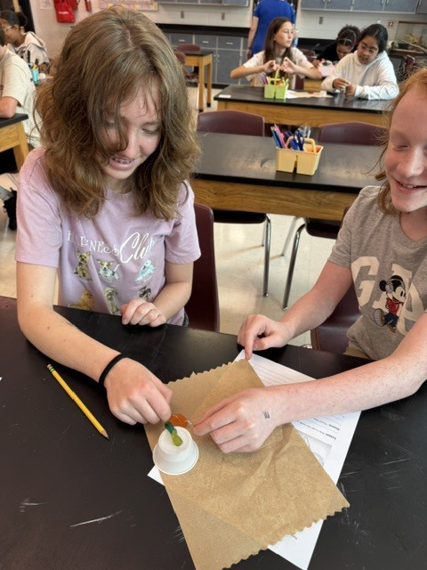 Two young girls are seated at a table, working on a project. One holds a pencil and a paper bag.