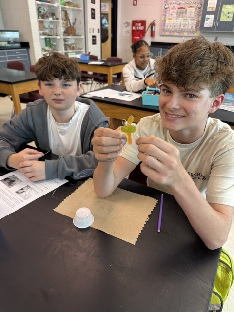 Two boys in a classroom with lab tables, one holding a small object, the other smiling and seated at a table with a piece of paper.