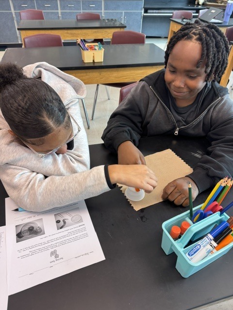 Two students at a desk working with craft materials and a worksheet. One student holds a glue stick.