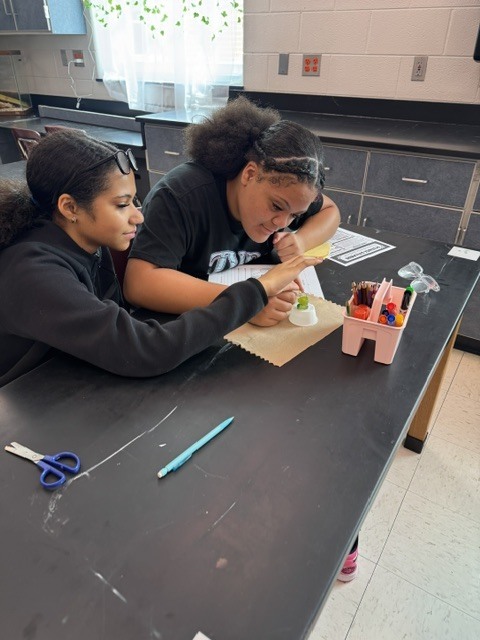 Two students work at a lab table, one holding scissors and the other a pencil, with art supplies.