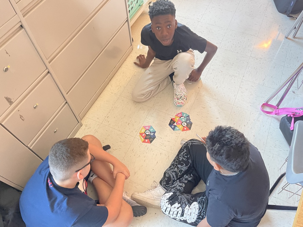 Three boys sitting on a floor, two wearing blue shirts. One boy is kneeling with a game board in front.