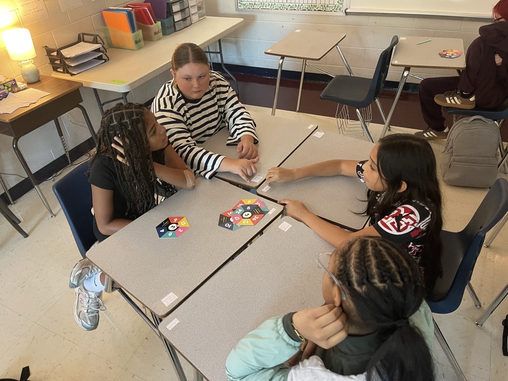 Students are seated at desks in a classroom, engaging with a teacher. Two students look at a game while another observes.