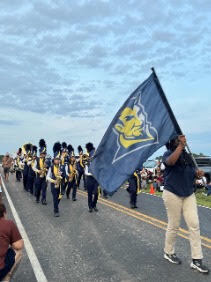 A marching band, carrying a blue flag, moves down a road. Spectators line the sides.