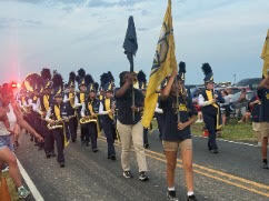 A marching band plays instruments and carries flags down a street. One person holds a large yellow flag.