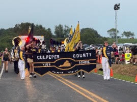 A school marching band holds a banner reading "Culpeper County High School" while walking down a road.