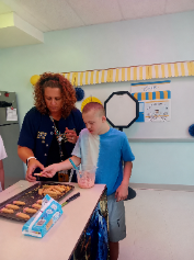 Adult and child stand at table, preparing food. Tray with cookies. Background has blue and white decorations.