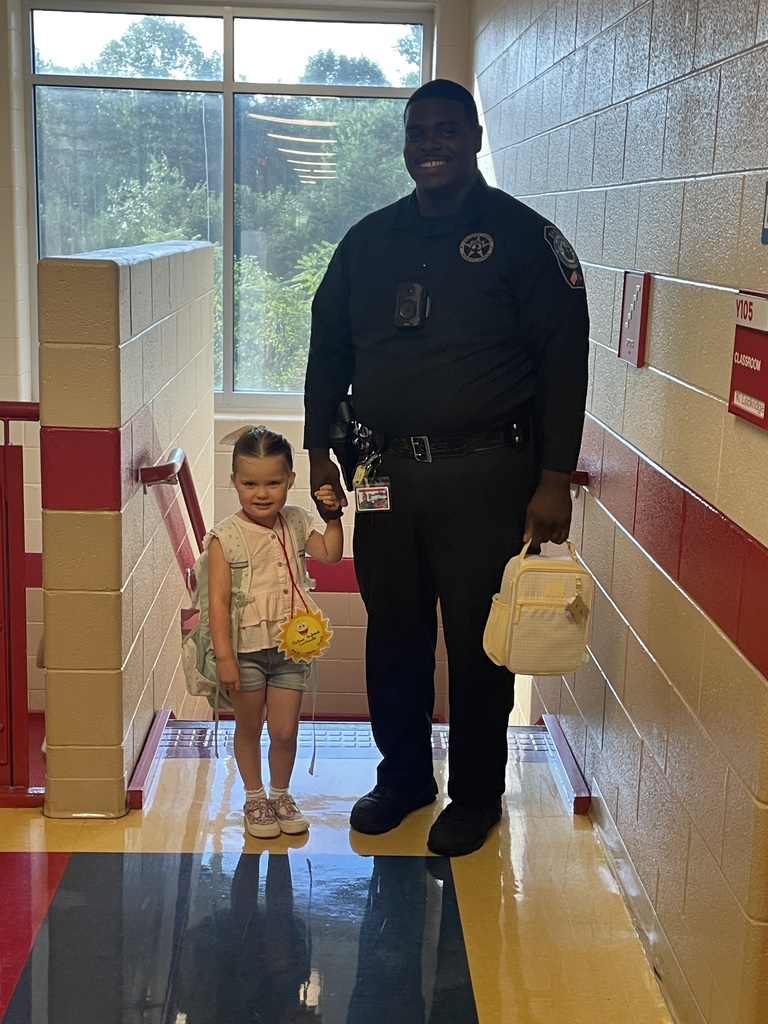 A police officer stands in a school hallway, holding a young child's hand and a bag, with a window behind them.