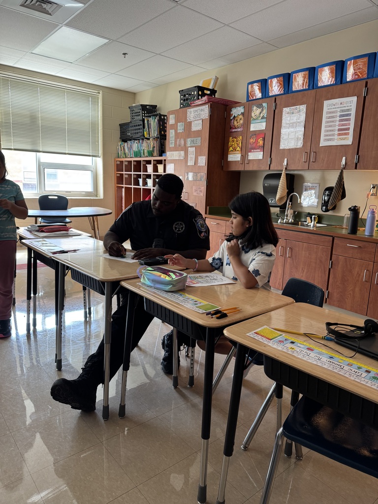 A police officer sits at a desk with a student in a classroom. Behind them, two people stand.