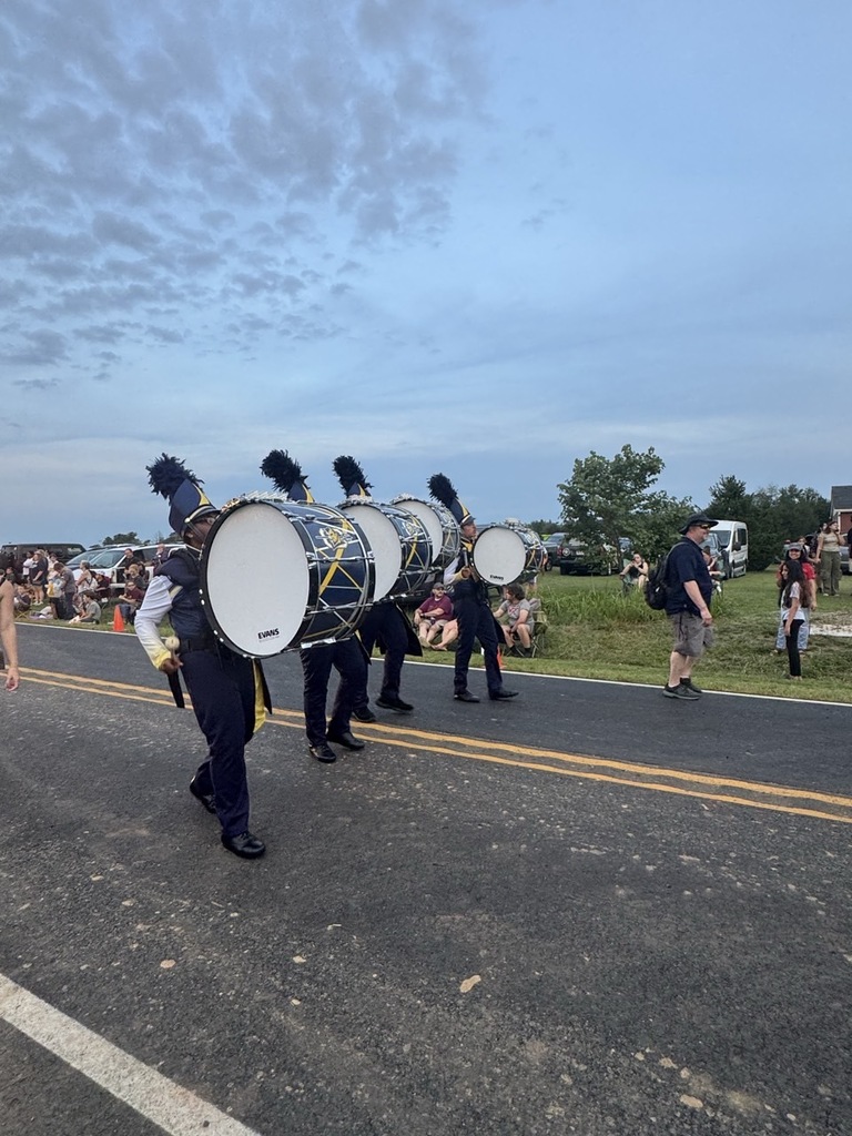 A marching band with drums on their shoulders is walking on a street. People are watching from the sides.