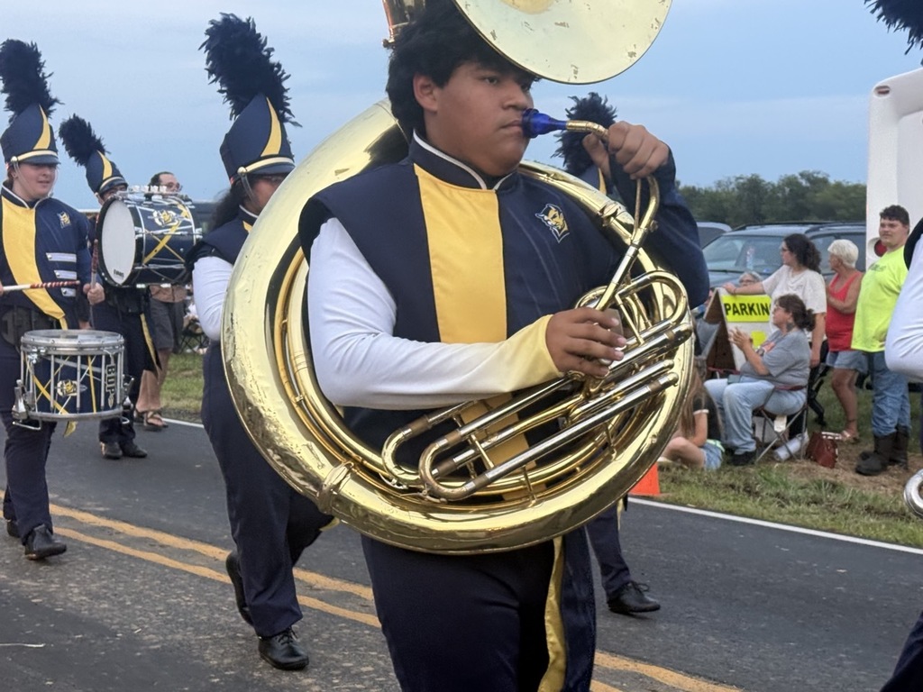 A marching band member plays a tuba while marching. Other members play drums. People watch from the roadside.