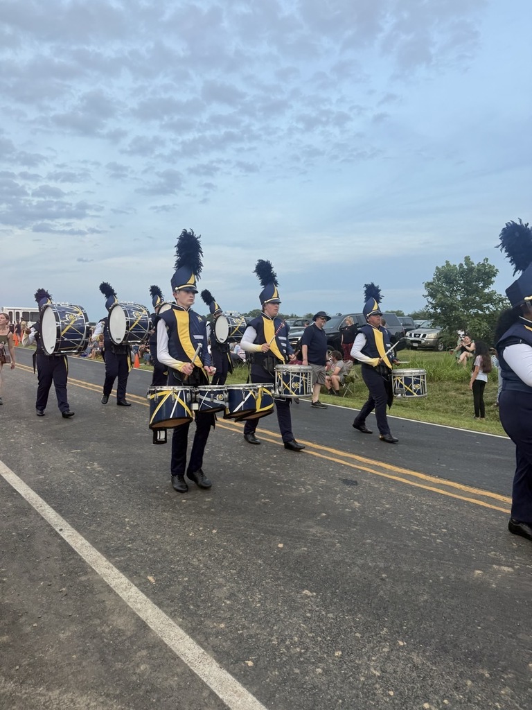 A marching band plays drums and other instruments down a road under a partly cloudy sky.