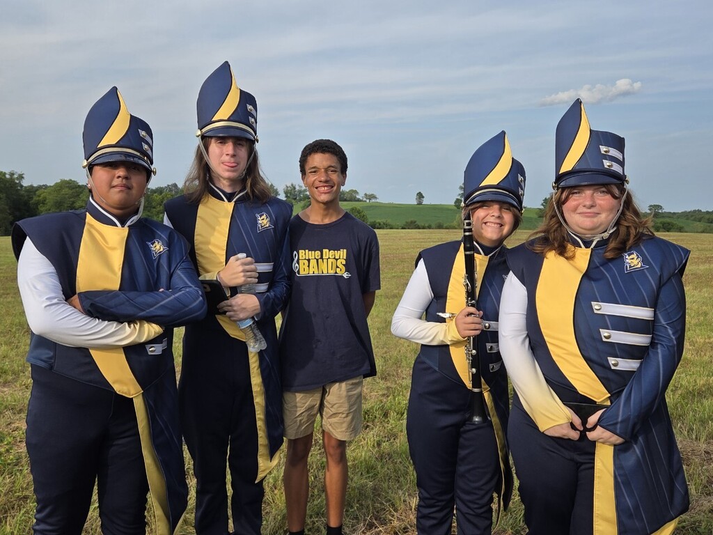 Five people in marching band uniforms stand in a grassy field with trees and blue sky.