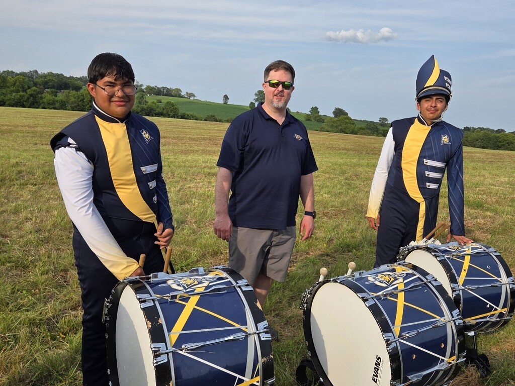 Three individuals stand in a field, two with drums and one without, wearing matching uniforms. One holds a drumstick.