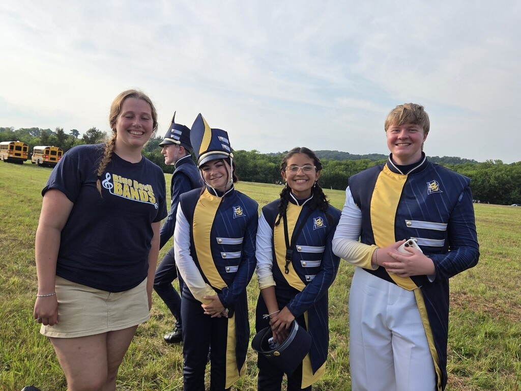 Four individuals in matching uniforms, two with hats, stand in a grassy field with a bus in the distance.