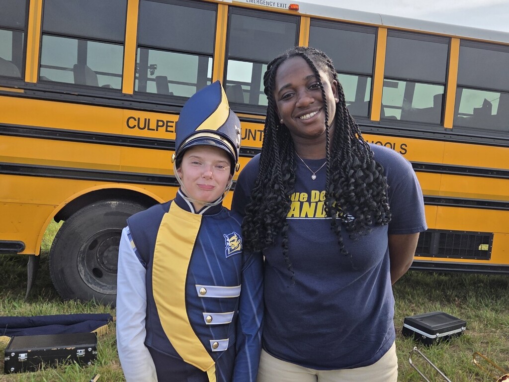 Two people stand outside; one wears a marching band uniform, the other a navy blue shirt. A yellow school bus is in the background.